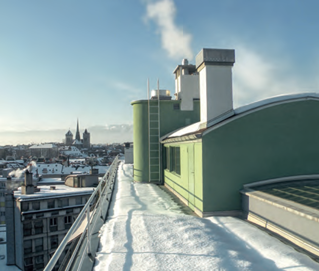 Verschneites Dach mit grünem Aufbau, Kaminrauch und Stadtsilhouette mit Kirchtürmen im Hintergrund